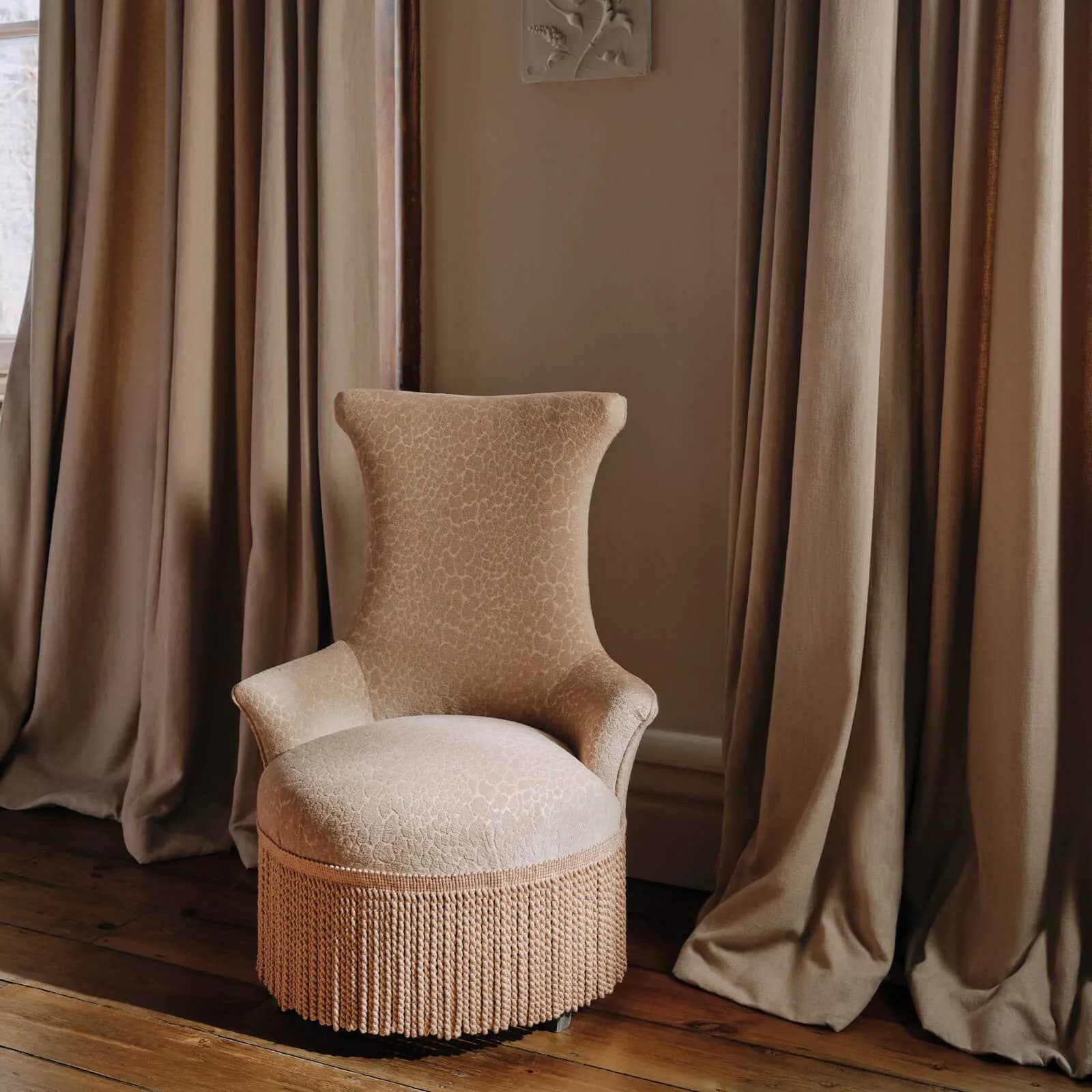 A softly upholstered accent chair with fringe trim framed by warm drapery.